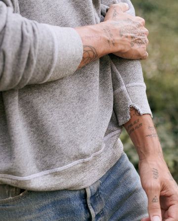 Close-up of a person wearing The Men's College Sweatshirt in Varsity Grey by THE GREAT. MAN, pulling up their sleeve to reveal tattoos on their hands and arms. Only the person's torso and arms are visible against a blurred outdoor background, giving a vintage-inspired vibe.