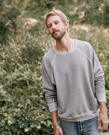 A bearded man with tattoos stands outdoors wearing THE GREAT. MAN’s Men’s College Sweatshirt in Varsity Grey and jeans. He is looking slightly to the side, exuding a comfortable vibe with greenery in the background.