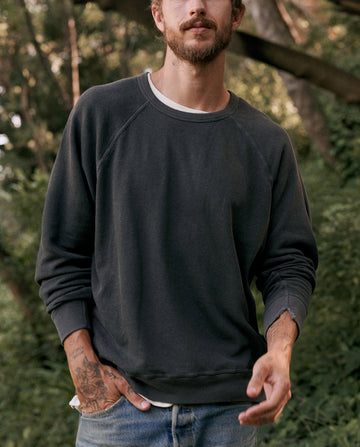 A person with a beard and tattoos on their hand is wearing The Men's College Sweatshirt in washed black by THE GREAT. MAN and jeans, standing in an outdoor setting with greenery in the background.