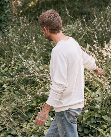 A person with short hair wearing The Men's College Sweatshirt in washed white by THE GREAT. MAN and blue jeans walks through a lush, green garden.