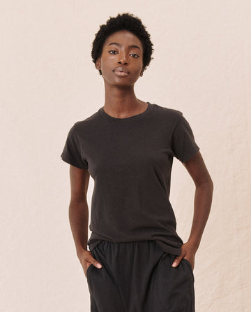 A person wearing THE GREAT. CORE KNITS' The Slim Tee in "Almost Black" and black pants stands against a plain light-colored background, with hands in pockets, looking directly at the camera.