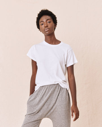 A person stands against a plain background, wearing The Slim Tee in True White by THE GREAT. CORE KNITS and grey pants, with their hands in their pockets.
