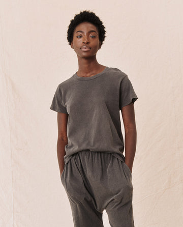 A person stands against a neutral background wearing The Slim Tee in Washed Black from THE GREAT. CORE KNITS and loose-fitting grey pants, with hands in pockets. The vintage tee, made of 100% cotton, complements the laid-back vibe perfectly.