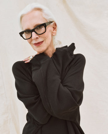 An older person with white hair, wearing black-framed glasses and The Slouch Sweatshirt in Solid -- Almost Black by THE GREAT. CORE KNITS, stands against a light background with a hand raised to their face.