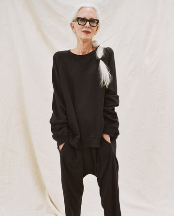 An older person with white hair tied in a side ponytail wears black glasses and "The Slouch Sweatshirt" in solid almost black from THE GREAT. CORE KNITS, against a plain beige background, with hands in pockets.