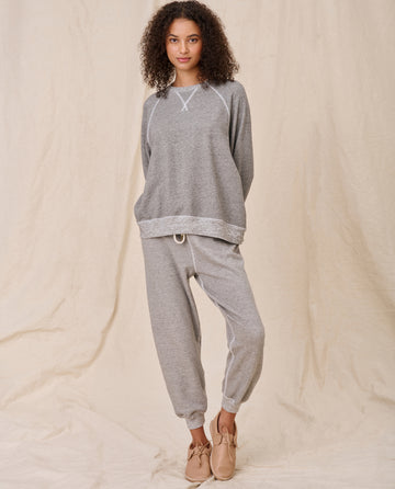 A person with curly hair wearing The Slouch Sweatshirt in Varsity Grey by THE GREAT. CORE KNITS, along with matching jogger pants, stands against a beige background. They are also wearing beige shoes.