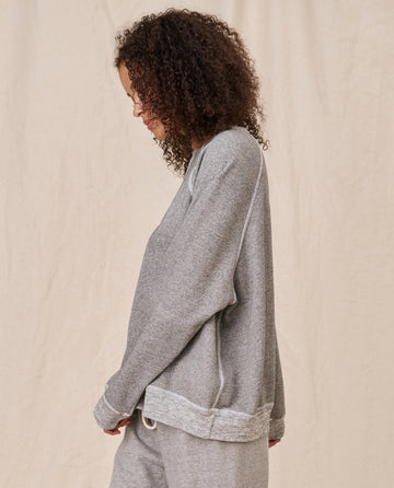 A person with curly hair, wearing The Slouch Sweatshirt in Varsity Grey from THE GREAT. CORE KNITS, with fuller sleeves and matching pants, stands in profile against a light beige background.