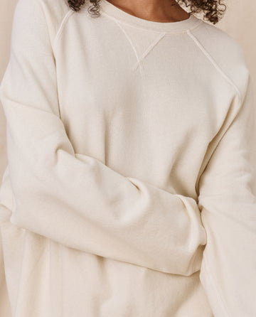 Person wearing The Slouch Sweatshirt in washed white, a product of THE GREAT. CORE KNITS. This oversized, vintage-feel, cream-colored sweater with long sleeves made from 100% cotton proudly carries the mark of American craftsmanship. Their torso and part of their arm are visible, with hands crossed at the waist.