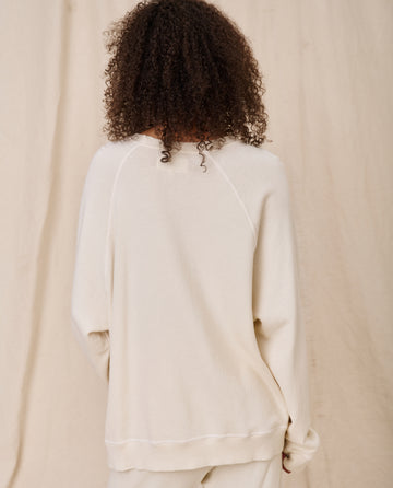 A person with curly hair is shown from the back, wearing The Slouch Sweatshirt in Washed White by THE GREAT. CORE KNITS, paired with 100% cotton vintage pants, standing in front of a beige fabric backdrop.