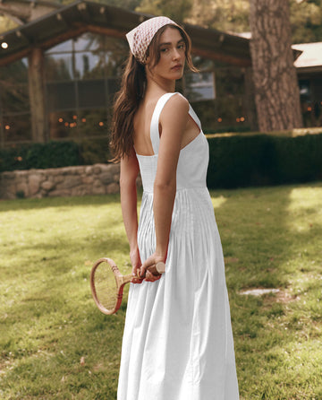 A woman in THE GREAT. SU25 Day Break Dress in white stands on grass, holding a wooden tennis racket behind her back, with a rustic building in the background, evoking timeless fashion charm.