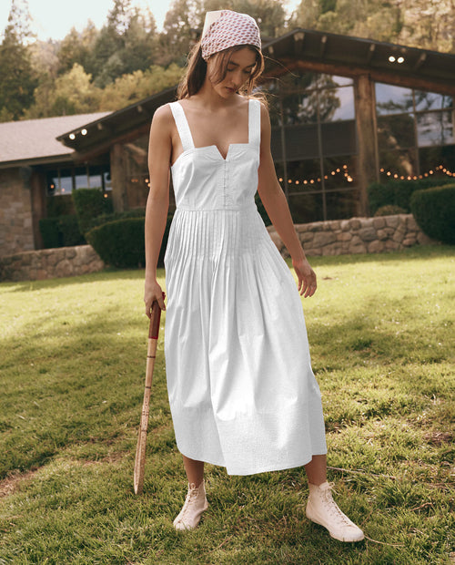 A woman in THE GREAT. SU25 Day Break Dress in White, paired with a headscarf and sneakers, stands on grass holding a wooden mallet, with a rustic building and trees in the background.
