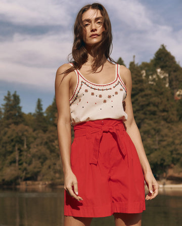A woman stands outdoors by trees and water wearing THE GREAT. SU25 The Beaded Slim Tank in Washed White, paired with high-waisted red shorts.