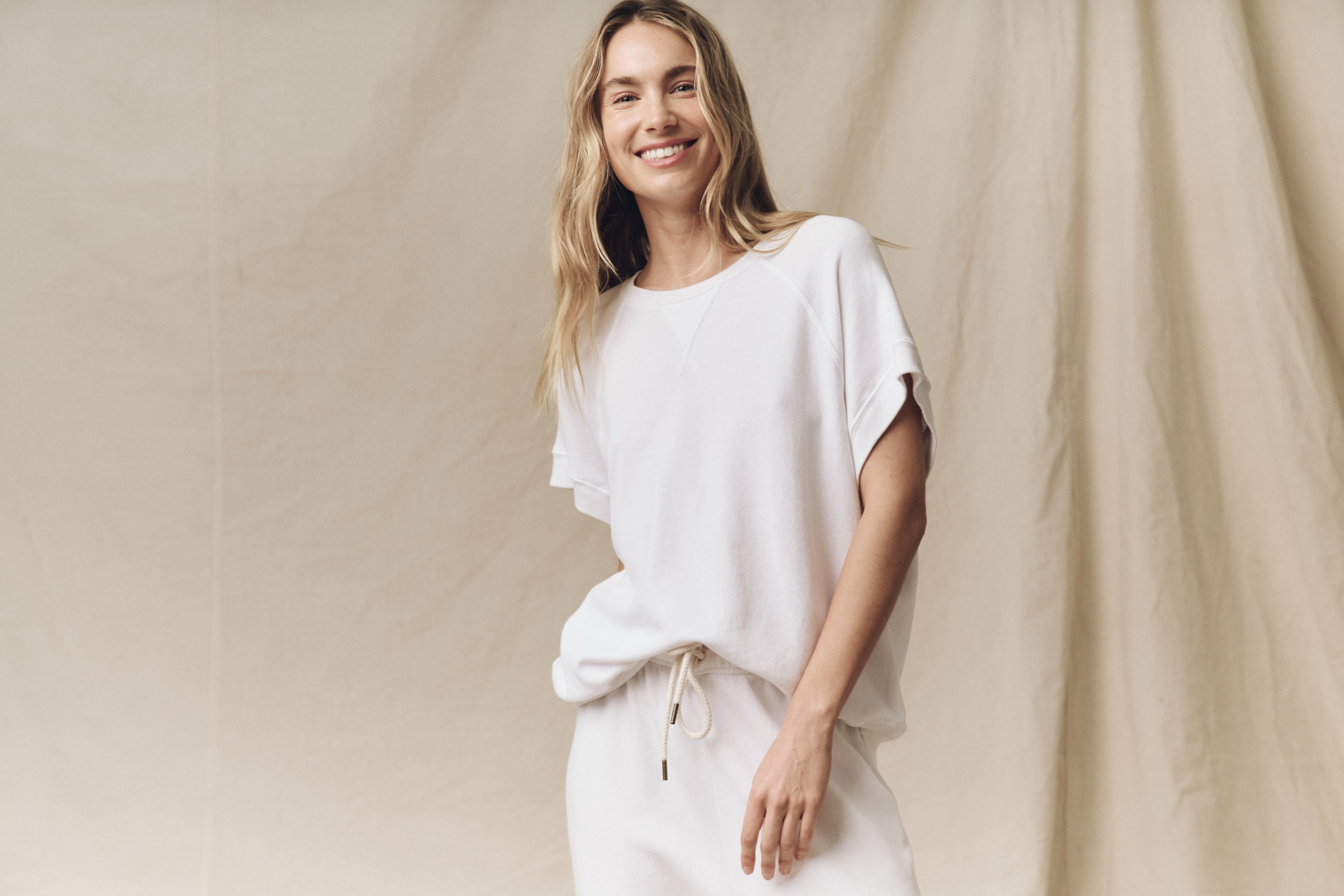 A person wearing a white outfit smiles while standing against a beige fabric background.