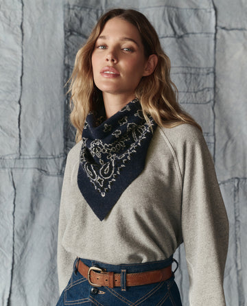 A woman wearing THE GREAT. H25 COLLECTION’s The Bandana Kerchief in Navy, a grey long-sleeve shirt, and high-waisted blue jeans stands before a textured fabric backdrop.