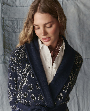 A woman with wavy hair looks down, wearing a white shirt, brown scarf, and THE GREAT. H25 COLLECTION's The Bandana Riding Cardigan in Navy, standing before a light fabric backdrop.