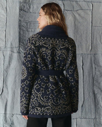 A person with long hair models THE GREAT. H25 COLLECTION's The Bandana Riding Cardigan in navy, featuring a belt and bandana pattern, viewed from the back against a textured gray wall.
