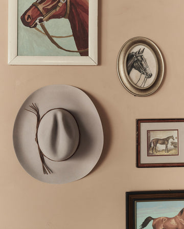 The Gus 6X Hat in Ranch Tan by THE GREAT. STETSON SP26 hangs on a beige wall, surrounded by framed paintings and illustrations of horses.