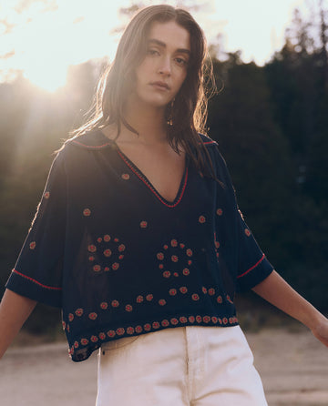 A woman with long hair wears THE GREAT. SU25's Beaded Vintage Sailor Top in navy and white pants, standing outdoors with sunlight behind her and trees in the background.