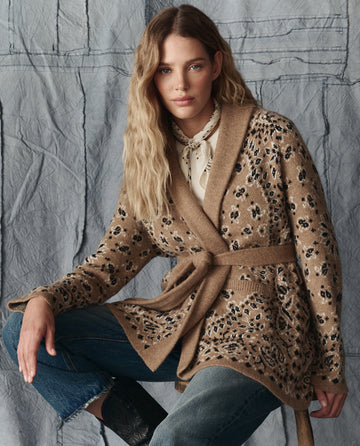 A woman with wavy blonde hair wears THE GREAT. H25 COLLECTION’s Bandana Riding Cardigan in Camel over a light blouse and blue jeans, seated before a draped gray fabric background.