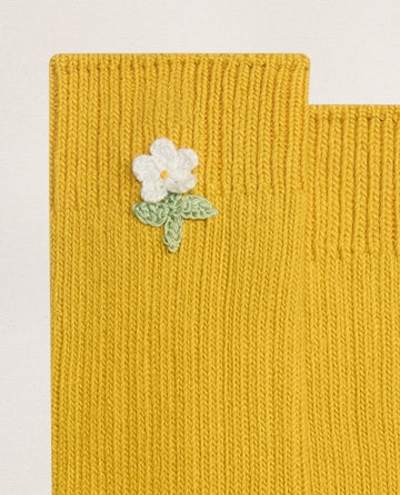 A close-up of EMME’s THE CROCHET FLOWER SOCK (1 FLOWER) -- MARIGOLD shows yellow knit cotton with a hand-crocheted white flower and green leaves near the top edge, adding vintage charm.