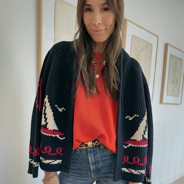 A woman wearing a navy cardigan with sailboat patterns, an orange shirt, and jeans stands indoors in front of framed artwork.