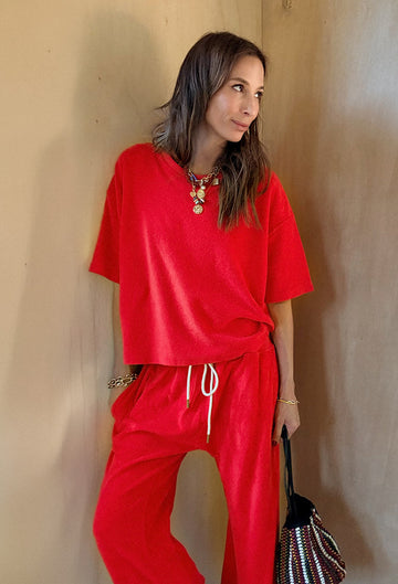 A woman stands indoors wearing a loose-fitting red top and matching red pants, accessorized with a colorful necklace and holding a beaded handbag.