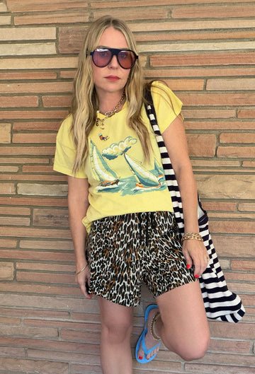 Woman wearing sunglasses, a yellow T-shirt with a sailboat graphic, leopard print shorts, blue sandals, and a striped bag stands against a brick wall.