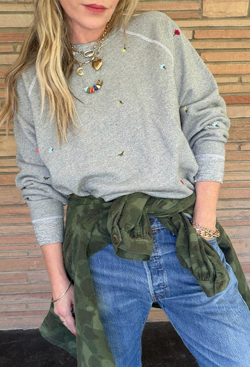 A person wearing a gray sweatshirt with small embroidered details, blue jeans, layered necklaces, and a green jacket tied around their waist, standing in front of a brick wall.