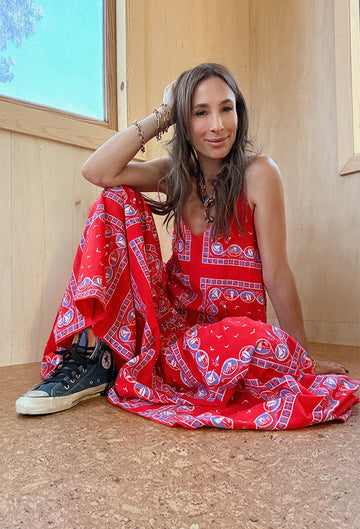A woman in a red patterned dress sits on a cork floor in a wooden room, with one leg bent and wearing black sneakers.