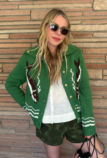 A person with long blonde hair wearing sunglasses, a green knit sweater with fish designs, a white shirt, and camouflage shorts stands in front of a brick wall.