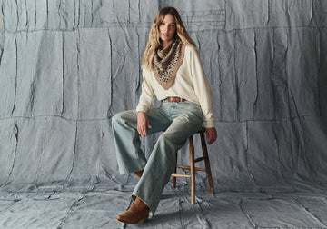 A woman sits on a wooden stool against a textured gray backdrop, wearing a cream sweater, wide-leg jeans, a patterned bandana, and brown boots.