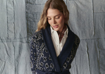 A woman with wavy hair looks down, wearing a white button-up shirt and a dark blue patterned cardigan, standing in front of a textured gray fabric backdrop.