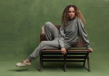 A person with curly hair wearing a grey sweatsuit and white sneakers sits on a wooden bench against a green background.