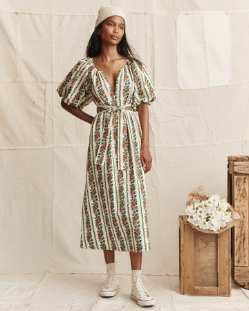 Woman wearing a floral striped dress, white knit hat, and white sneakers stands indoors next to wooden crates and a bouquet of white flowers.