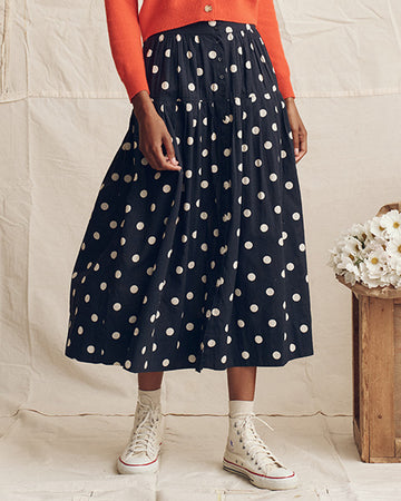 Person wearing a navy blue skirt with white polka dots, an orange top, and white high-top sneakers, standing next to a wooden crate with white flowers.