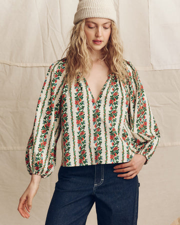 A person wearing a knitted beanie, a floral patterned blouse with balloon sleeves, and dark jeans stands against a neutral fabric backdrop.