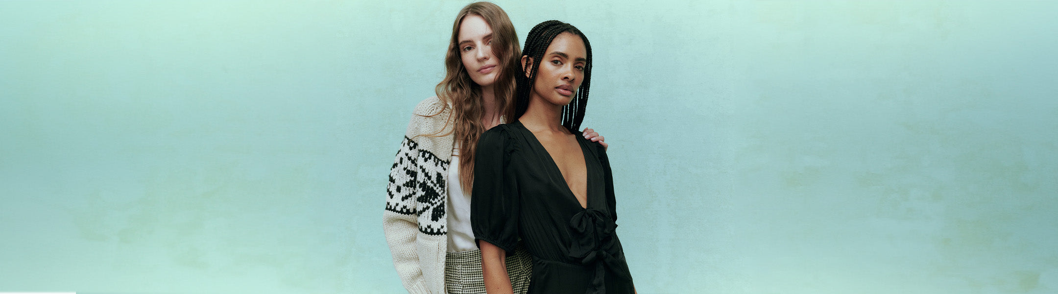 Two women standing side by side, one wearing a patterned sweater and the other in a black dress, against a soft green background.