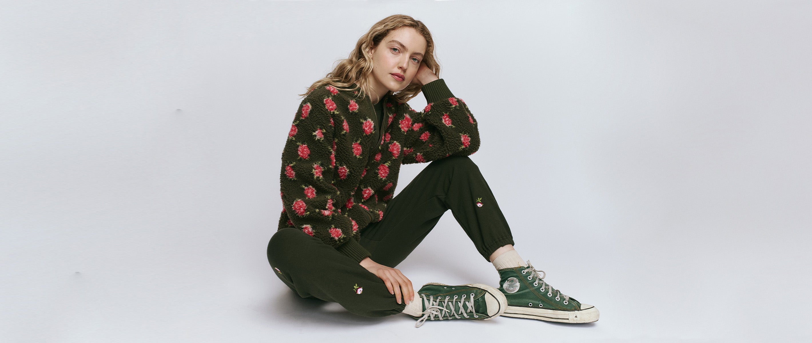 A person sits on the floor against a plain background, wearing a floral-patterned sweater, dark pants, and green high-top sneakers.
