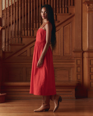 A woman stands indoors on a wooden floor, wearing a red sleeveless dress and tan cowboy boots, with long hair draped over her shoulder; a wooden staircase is in the background.