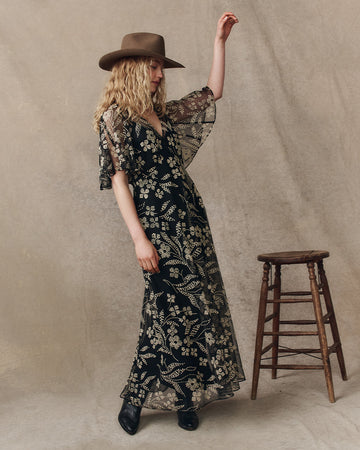 A woman in a black floral dress and brown hat stands next to a wooden stool against a neutral backdrop.