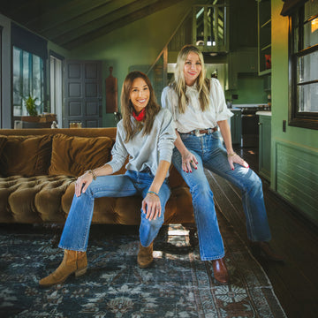 Two women in casual jeans and boots pose together in a green living room with a brown velvet sofa and a patterned rug.