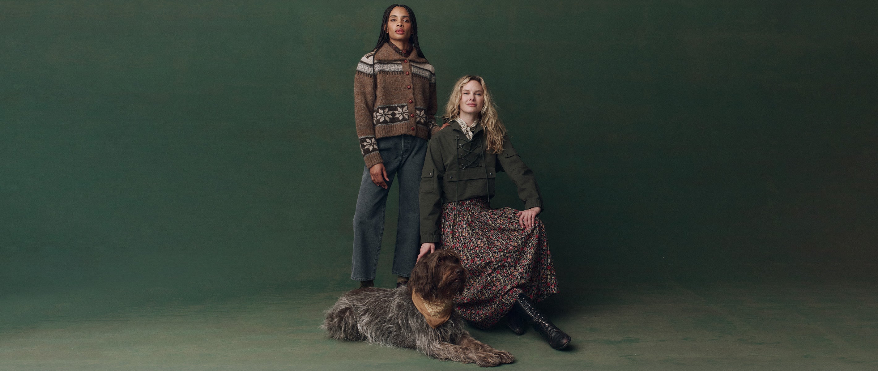 Two women pose in front of a dark green backdrop; one stands wearing a patterned sweater, the other sits in a floral skirt and jacket, with a large shaggy dog lying in front of them.