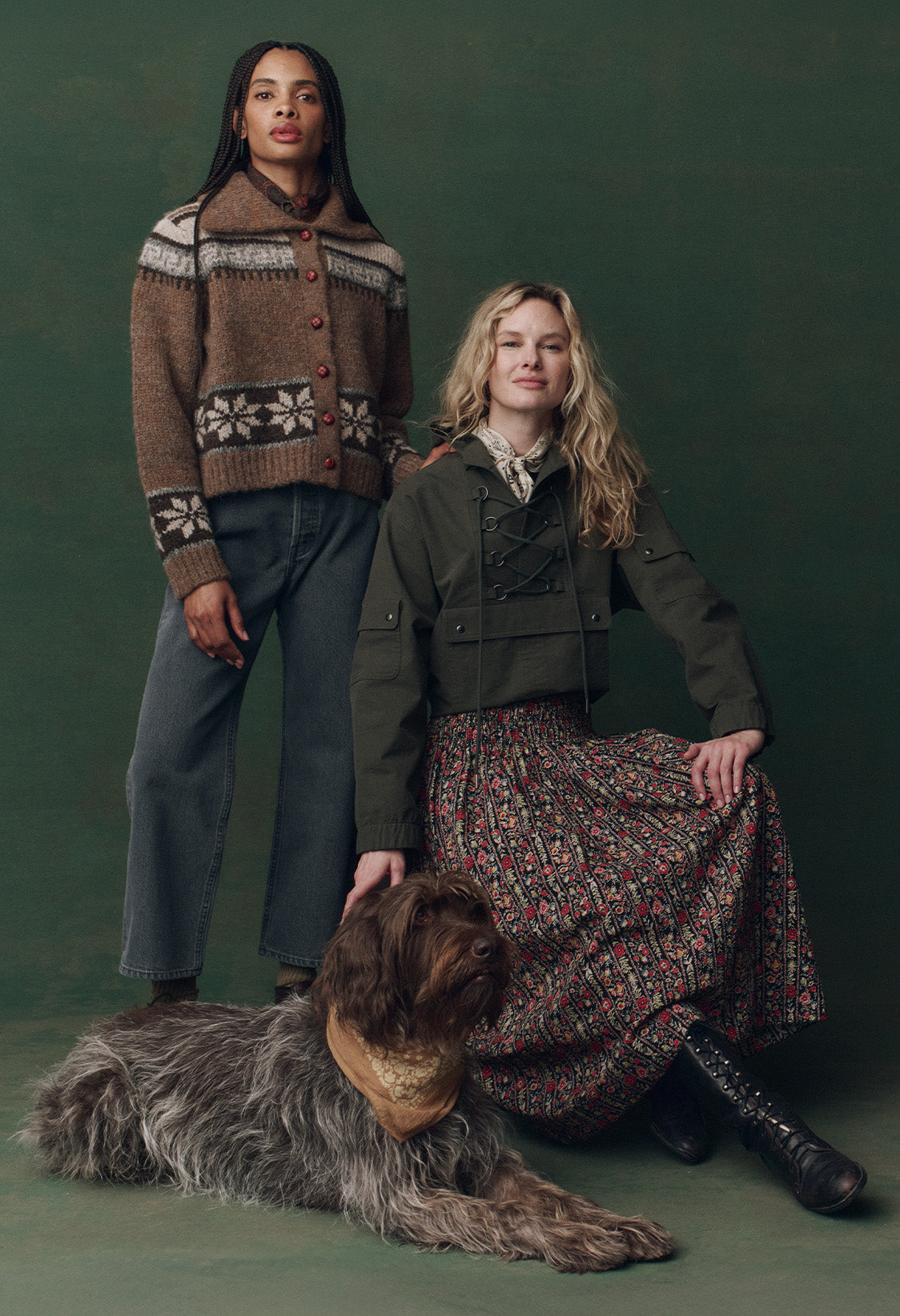 Two women pose against a green backdrop; one stands in a patterned sweater and jeans, the other sits in a green jacket and floral skirt with a large, shaggy dog at her feet.