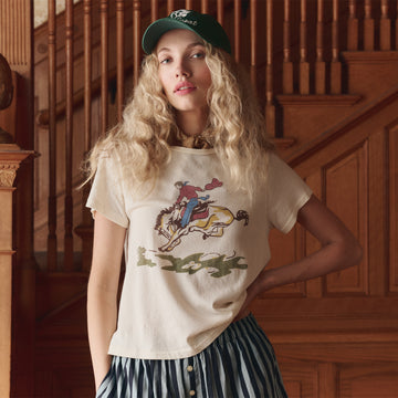 A person with long curly blonde hair wearing a white t-shirt with a horse graphic, a green cap, and a striped skirt stands in front of wooden stairs.