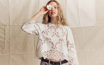 Woman with wavy hair wearing a white lace blouse and jeans holds a white flower over her eye, standing in front of a beige fabric backdrop.