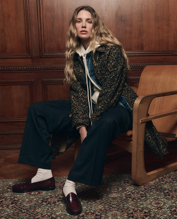 A woman with long wavy hair sits on a wooden chair in a wood-paneled room, showcasing women's fashion. She wears THE GREAT. F26 COLLECTORS' The Trench Coat in Dark Leopard—a modern take on classic outerwear—styled with layered clothing, wide-leg pants, white socks, and burgundy loafers.