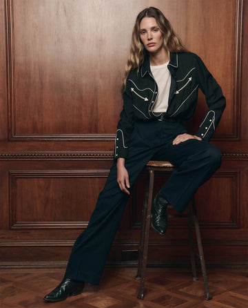 A person with long hair sits on a wooden stool, wearing THE GREAT. F26 COLLECTORS The Western Shirt in black over a white t-shirt, paired with dark pants and black boots, set against a brown wood-paneled wall.