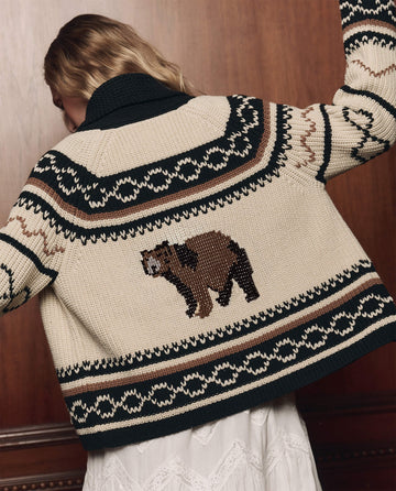 A person facing away, wearing The Bear Lodge Cardigan in black by THE GREAT. F26 COLLECTORS, which features dark green and brown patterns and a knit bear design on the back, standing in front of a wooden wall.
