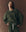 A person wears THE GREAT. F25 D1 The Slouch Sweatshirt. Novelty -- Forest Camo, paired with matching sweatpants featuring a white drawstring, and stands against a brown backdrop.