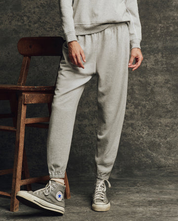 A person wearing THE GREAT. H25 CORE FLEECEBACK The Fleece Stadium Sweatpant in Heather Grey stands by a wooden chair against a textured dark background.
